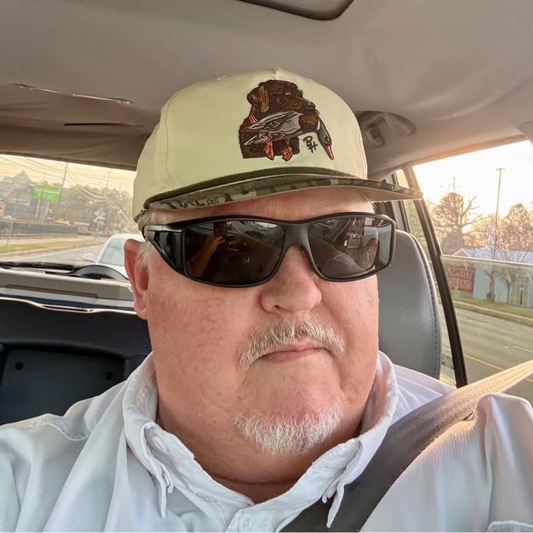 Man with mustache wearing cream-colored baseball cap and dark sunglasses, photographed inside a vehicle
